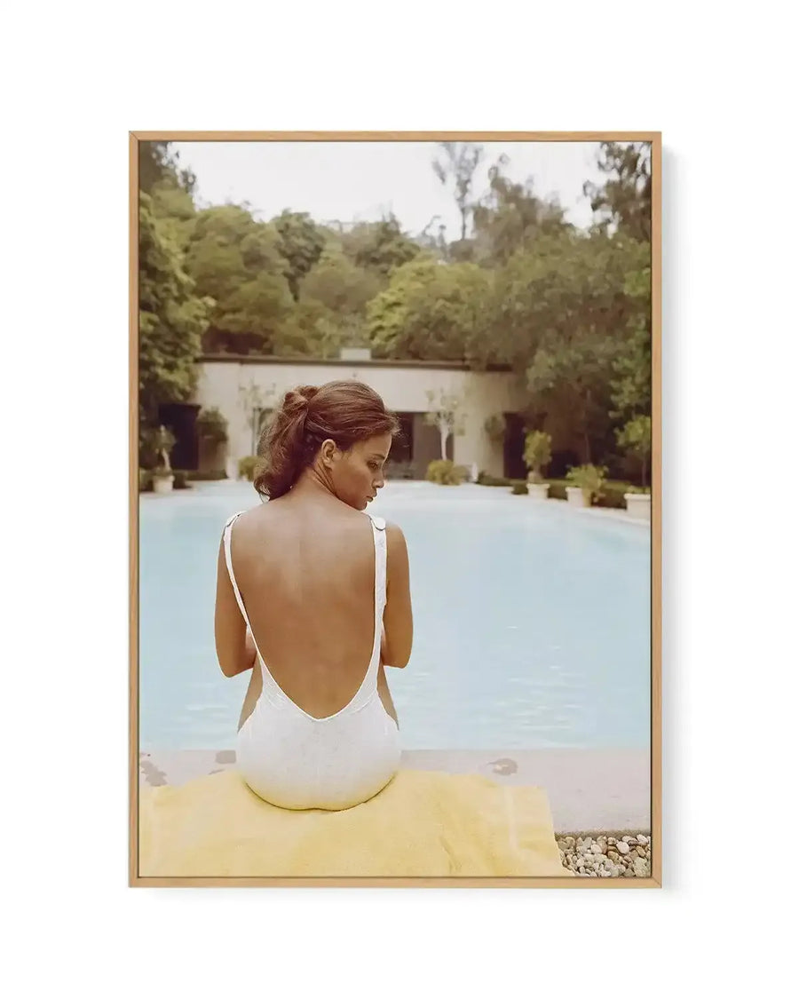 Framed canvas art print titled "Fille de Citron" featuring a photograph of a woman viewed from behind sitting on a yellow towel beside a pale blue pool. She wears a white backless swimsuit and has her reddish-brown hair styled in an updo. The scene is set in a Mediterranean-style courtyard with a white building, tall green trees, and landscaping. The photograph has warm, muted tones with a vintage quality. The canvas is mounted in a natural wood frame with white border.