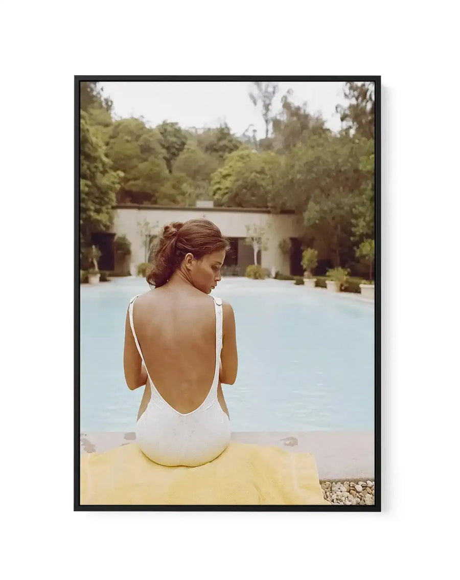 Framed canvas art print featuring a color photograph of a woman sitting on a yellow towel by a swimming pool, viewed from behind. She wears a white backless swimsuit and has her hair styled in an updo. The scene shows a serene courtyard setting with a light blue pool, cream-colored building with arched features, and dense green trees in the background under an overcast sky. The photograph has a soft, vintage color palette with warm and cool tones. The canvas is mounted in a black frame with a white border.