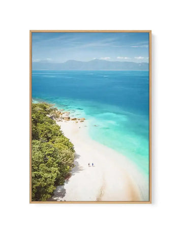 Framed canvas art print of Fitzroy Island, Queensland featuring an aerial view of a pristine white sand beach bordered by lush green tropical vegetation on the left. Crystal clear turquoise and aquamarine waters gradient to deeper blue in the distance, with granite rock formations visible along the shoreline. Mountains visible across the water under a bright blue sky with wispy clouds. The photograph is mounted in a natural wood frame with a white border.