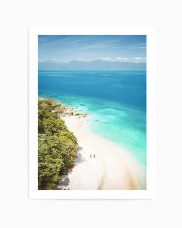 Framed aerial photograph of a tropical beach with a natural wood frame and white border. The composition features a long stretch of white sand curving along the coastline, bordered by lush green trees on the left and clear turquoise ocean on the right. Two small figures walk on the sand, with distant mountains visible on the horizon under a bright blue sky.