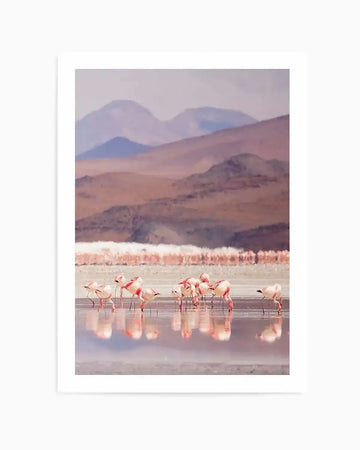 Art print featuring a serene landscape with a flock of flamingos wading in a shallow lake, set against a backdrop of layered mountains under a soft, muted sky. The flamingos display shades of pale pink and white, with their reflections visible in the still water. The mountains in the distance are rendered in soft purples, browns, and dusty rose hues, suggesting a hazy, distant atmosphere. The overall impression is a tranquil, painterly scene with a gentle colour palette.