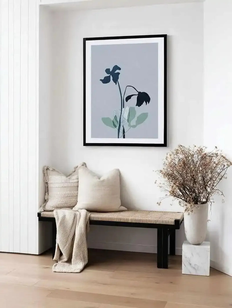 Framed art print titled Fleur Graphique II displaying two wilting flowers with dark blue-black petals and green leaves against a soft blue-grey background. Black wooden frame with white border. Print shown hanging on white paneled wall above a black bench with woven seat, cream cushions, and a cream throw blanket. White ceramic vase with dried baby's breath sits beside the bench on a marble block. Natural wood flooring visible in foreground.