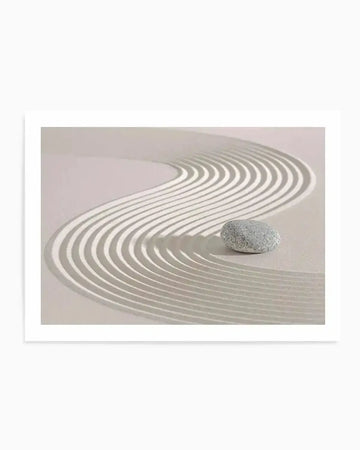 Framed photograph of a minimalist zen garden with a single grey stone on raked sand, with a white frame and white border. The sand is a pale beige, and the raked lines form a graceful, undulating S-curve across the frame, creating shadows that add depth and texture to the composition.