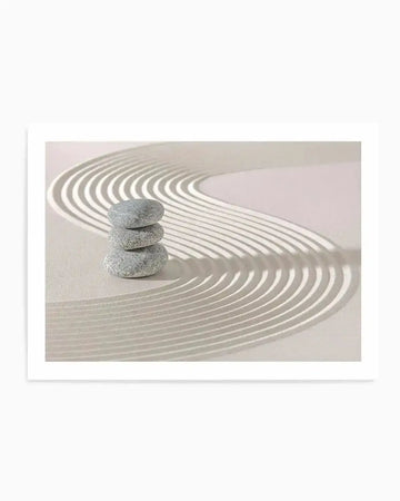 "Art print featuring a close-up photograph of a zen garden with raked sand and a stack of three smooth grey stones. The sand is a pale cream colour with curved, parallel lines raked into it, creating a wavy pattern that suggests ripples or water. The stones are stacked vertically in the lower left quadrant of the image, casting soft shadows. The overall mood is calm and minimalist."