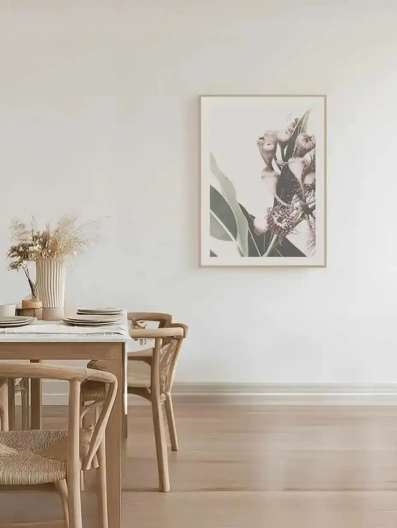 Lifestyle shot of a minimalist dining room featuring a framed art print of flowering gum. The print, displayed on a white wall, shows close-up eucalyptus flowers and gum nuts in soft pinks and muted greens against a light background, with a natural wood frame and white border. A light wood dining table with woven chairs and a vase of dried grasses is in the foreground.