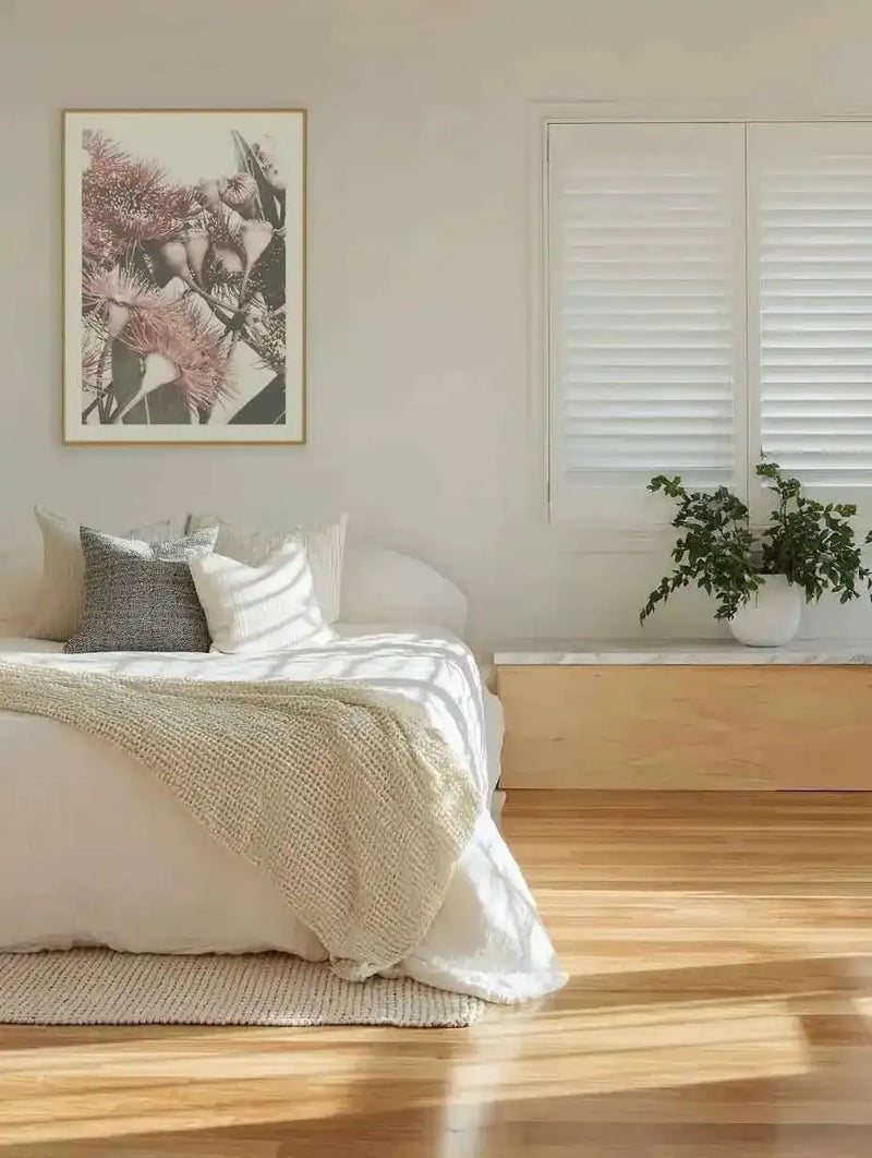 Flowering Gum II art print displayed on a cream wall above a bed with white pillows and natural linen bedding. The framed print features botanical illustration of dusty rose and mauve gum flowers with dark green foliage against a soft neutral background, presented in a natural wood frame with white border. Bedroom setting includes wooden flooring, white plantation shutters, potted green plant on windowsill, and neutral textured decor.