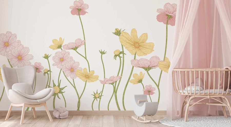 Lifestyle shot of a nursery with a peel and stick wallpaper mural featuring hand-drawn cosmos flowers in various shades of pink and yellow on a white background. The flowers are depicted with long, slender green stems and some show buds. On the left, a light grey upholstered armchair with light wood legs sits on a light wood floor. On the right, a light wood crib with a pink canopy is placed on a round, textured white rug.