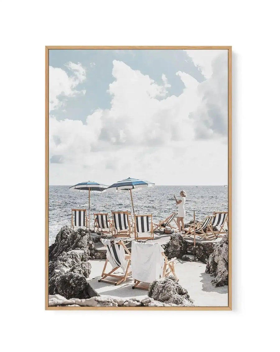 Framed canvas art print titled "Fontelina Days, Capri" featuring a scenic beach photograph. The image shows a sandy cove with rocky outcroppings, multiple striped deck chairs in navy and white, two open umbrellas, and a woman in a white dress standing near the water's edge. The sea is calm and grey-blue, with white clouds dotting a pale blue sky. The photograph has a warm, vintage-inspired aesthetic with natural lighting. The canvas is mounted in a natural wood frame with a white border.