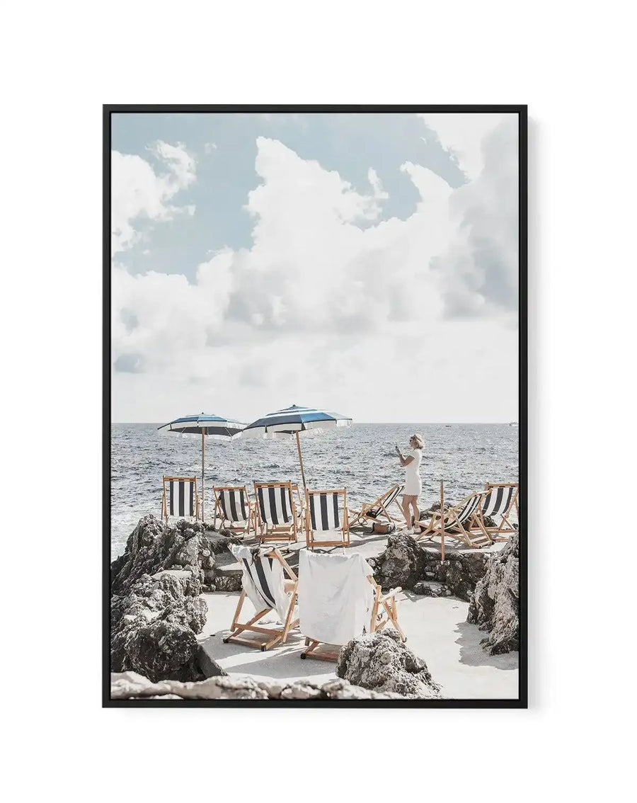 Framed canvas art print titled Fontelina Days, Capri featuring a serene beach scene with wooden striped deck chairs and blue umbrellas arranged on pale sand between rocky outcrops. A woman in a white dress stands in the distance by the turquoise sea. Soft white clouds fill a pale blue sky. The photograph has a calm, leisurely aesthetic with natural lighting and muted tones. Black frame with white border.