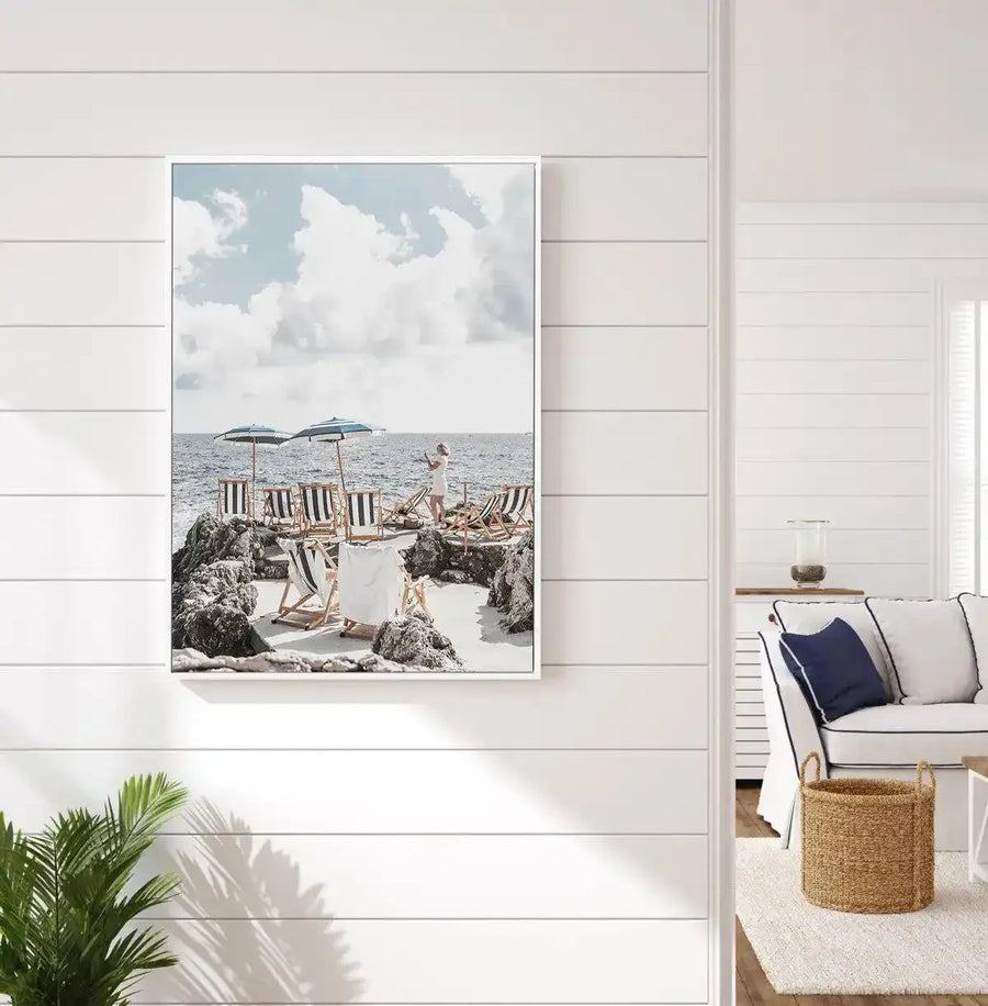 Lifestyle shot of a bright, coastal-style living room with a framed canvas print of a beach scene displayed on a white shiplap wall. The print features a rocky cove with white sand, two striped beach umbrellas, and several striped lounge chairs. A person in a white dress stands near the water. The room includes a white sofa with navy blue cushions, a woven basket, and a green plant in the foreground.
