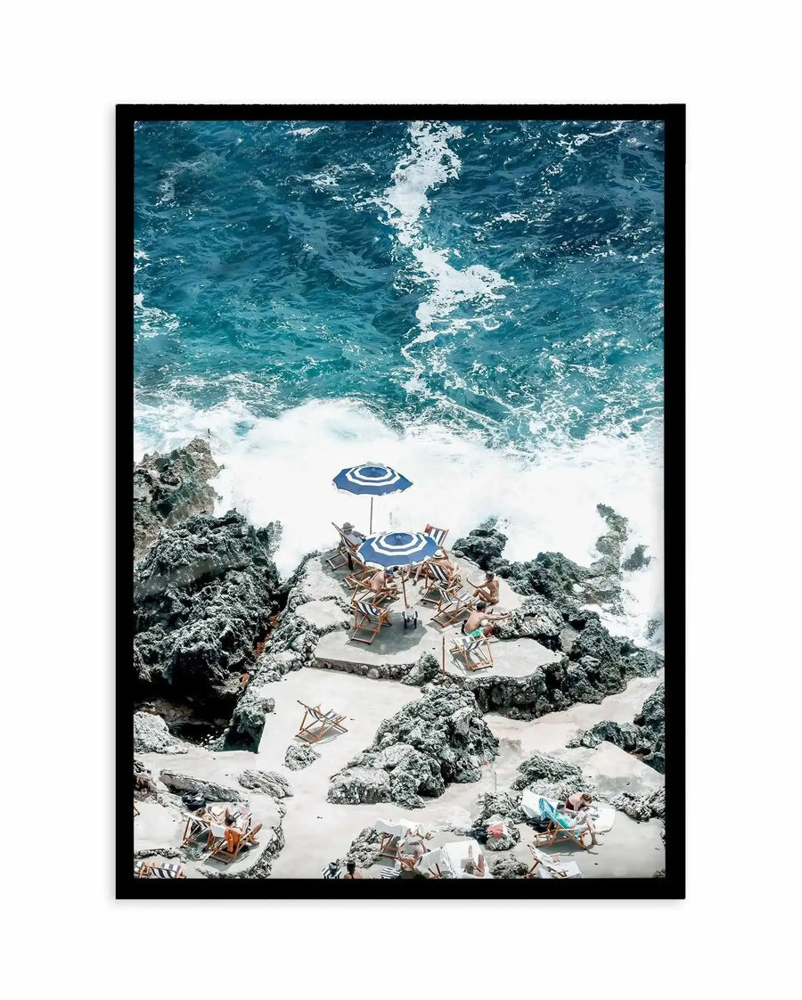 Framed art print featuring an aerial photograph of a sandy beach cove surrounded by rocky outcrops and turquoise ocean waves. Multiple people in beach chairs are scattered across the pale sand, with two blue and white striped umbrellas providing shade in the center. The composition captures the contrast between the white foam of breaking waves, the deep teal water, grey-green rocks, and light beige sand. The photograph is mounted in a black frame with a white border.