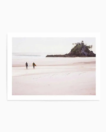 Framed photograph of a coastal beach scene with two surfers walking along wet, pale pink sand at low tide. Gentle waves break in the distance under an overcast sky, and a small, rocky island with trees and a wooden staircase is visible on the right. The print has a white border.