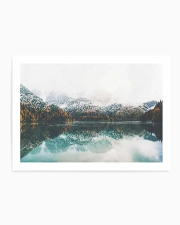 Framed photograph of a serene mountain lake reflecting snow-dusted autumn trees and an overcast sky, with a white frame and white border. The composition features a still body of water in the foreground, mirroring the snow-capped mountains and evergreen and deciduous trees with vibrant orange and yellow foliage lining the distant shore. The sky is a soft, hazy white, creating a tranquil and atmospheric scene.