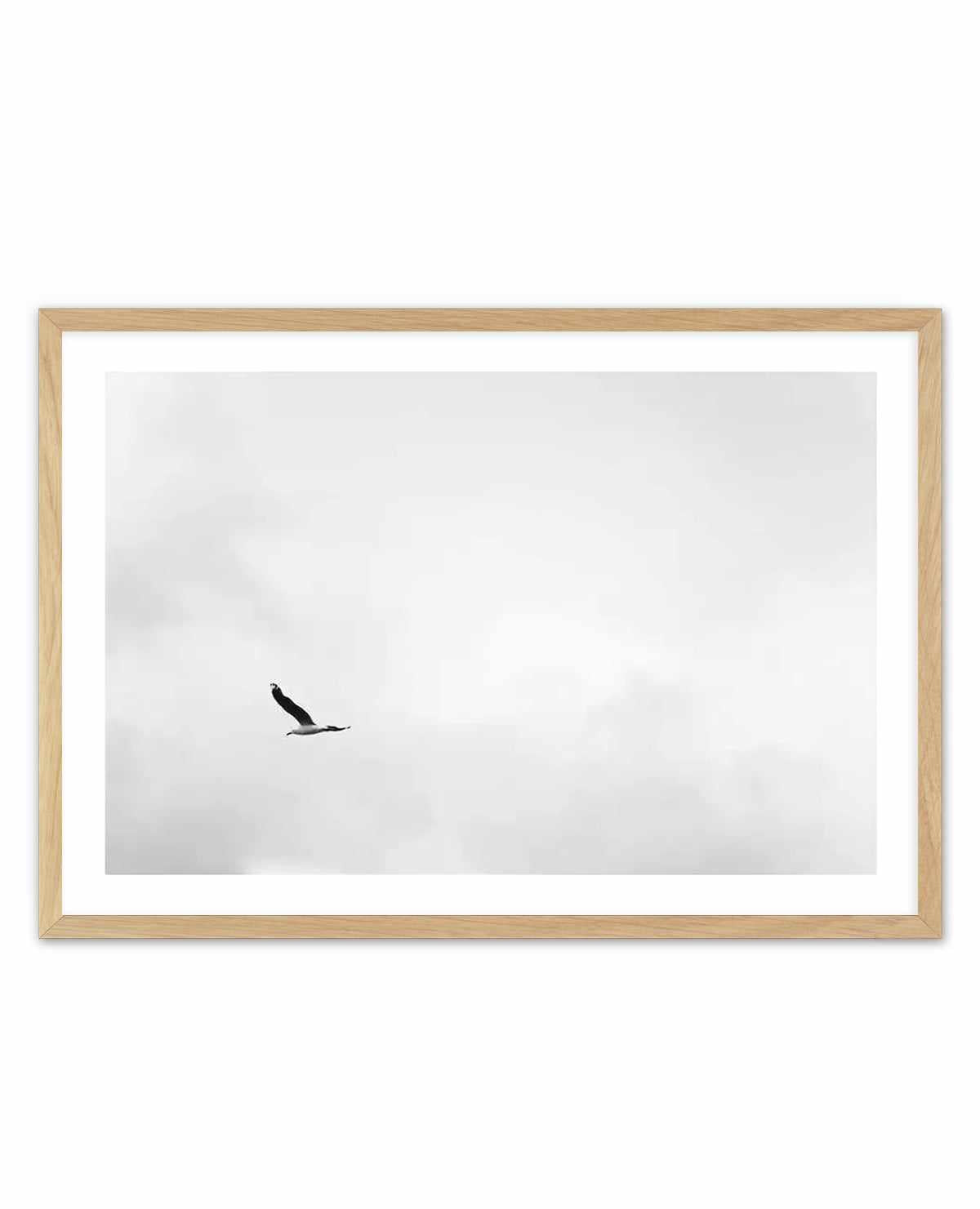 Black and white photograph of a single bird in flight against a light grey, cloudy sky, displayed in a natural wood frame with white border matting. The minimalist composition captures the bird mid-flight in the left-center area, creating a sense of solitude and freedom against the expansive, overcast sky. The photograph has a serene, contemplative quality with soft tonal gradations in the sky.