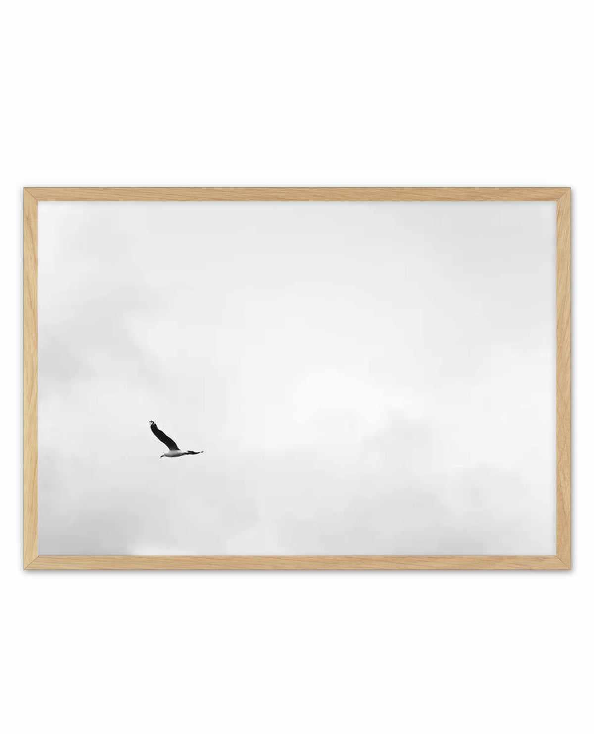Framed art print with natural wood border featuring a minimalist black silhouette of an eagle in flight against a cloudy white sky. The photograph-style image captures the bird mid-soar in the center-left portion of the composition, conveying a sense of freedom and solitude. The simple, high-contrast composition emphasizes negative space with soft gray and white tones throughout the overcast sky background.