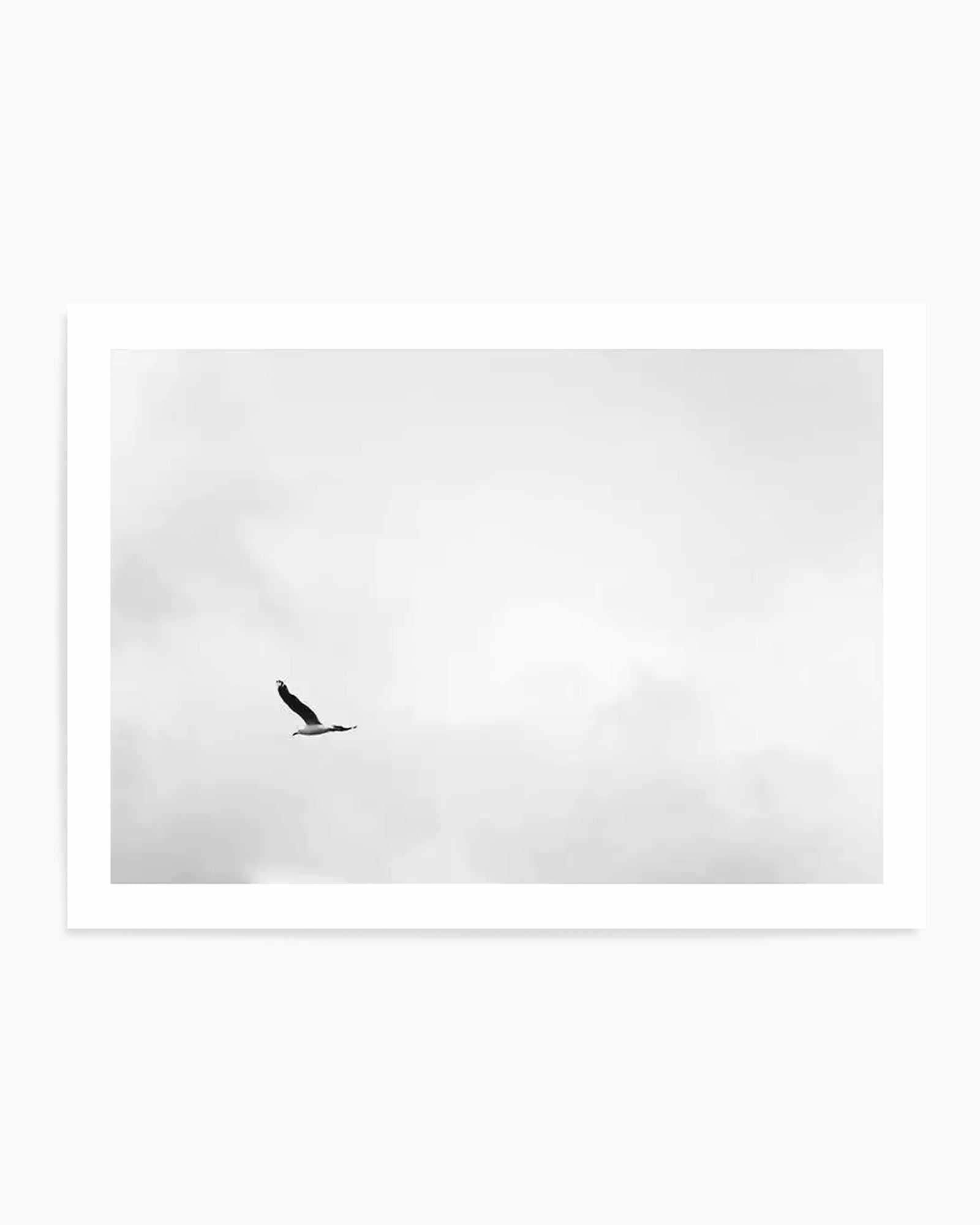 Black and white minimalist art print featuring a single bird in flight against a cloudy sky, captured in a stark, high-contrast photographic style. The bird is positioned off-center in the composition, silhouetted against soft, overcast clouds that fill most of the frame. The print is mounted in a white frame with a clean white border, creating a simple, serene aesthetic with ample negative space that emphasizes solitude and freedom.