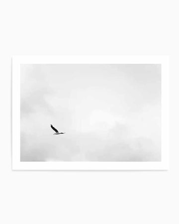 Black and white minimalist art print featuring a single bird in flight against a cloudy sky, captured in a stark, high-contrast photographic style. The bird is positioned off-center in the composition, silhouetted against soft, overcast clouds that fill most of the frame. The print is mounted in a white frame with a clean white border, creating a simple, serene aesthetic with ample negative space that emphasizes solitude and freedom.