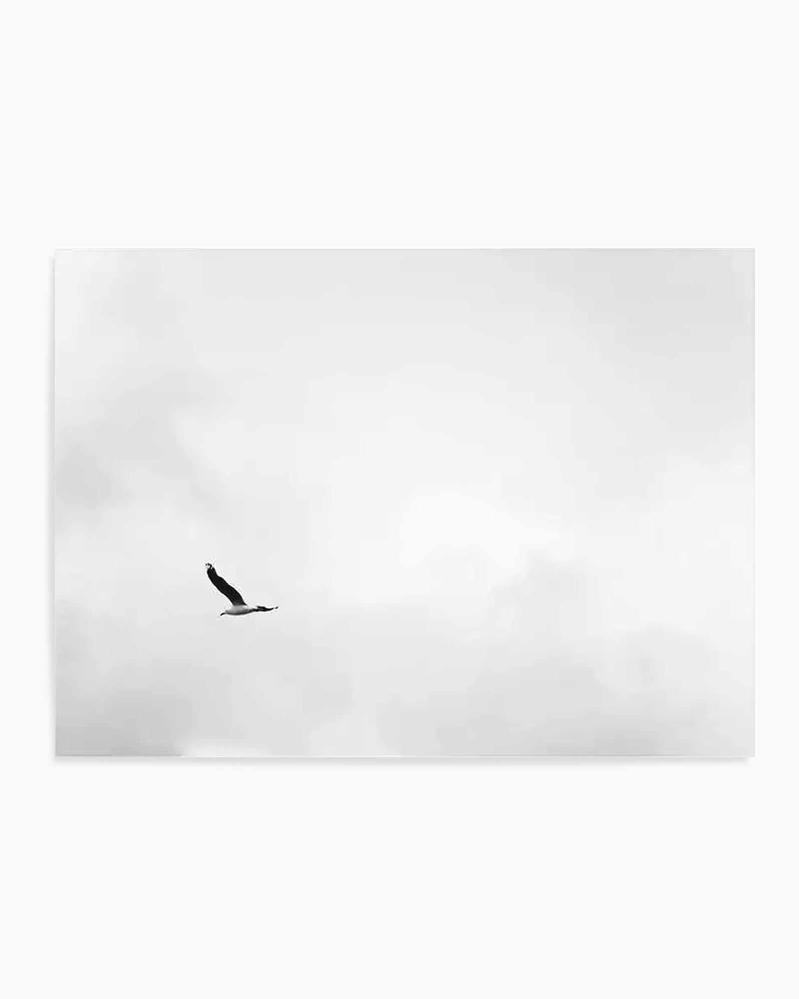 Minimalist black and white art print featuring a single bird in silhouette soaring through a cloudy, overcast sky. The bird appears to be an eagle or large raptor with wings spread wide, positioned slightly left of center against a soft, hazy grey and white gradient background with subtle cloud formations. The composition emphasizes negative space and simplicity, creating a serene, contemplative mood. The print has a clean white border and matte finish.