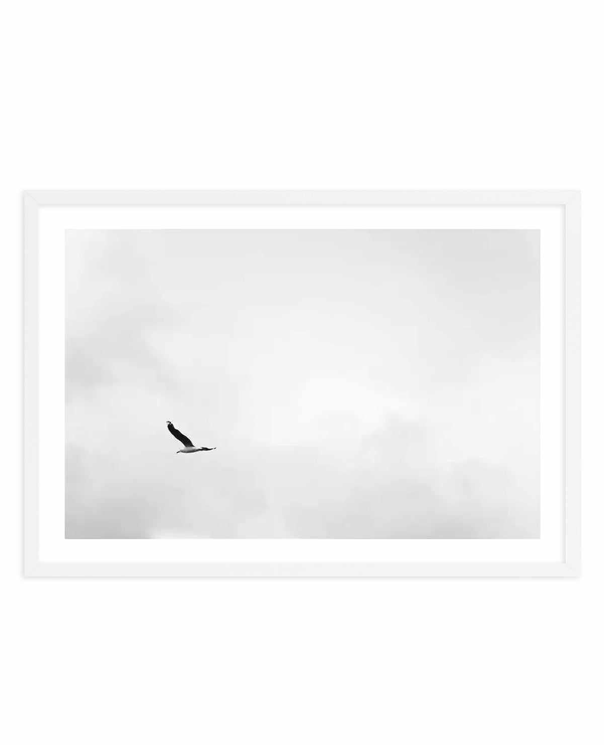 Black and white minimalist photograph of a single bird in flight against a overcast, cloudy sky. The bird appears small in the composition, positioned slightly left of center, creating a sense of solitude and freedom. The image has a grainy, soft quality typical of fine art photography. The artwork is presented in a white frame with a white border, displayed against a white background.