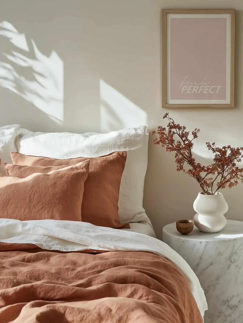 Framed art print with dusty rose background displayed above a marble nightstand in a minimalist bedroom. The print features lowercase text reading "fuck PERFECT" in light pink lettering, mounted in a natural wood frame with white border. The bedroom scene shows a bed with terracotta and cream linen bedding, white pillows, dried burgundy botanicals in a white ceramic vase, and natural sunlight casting shadows on the neutral beige wall.