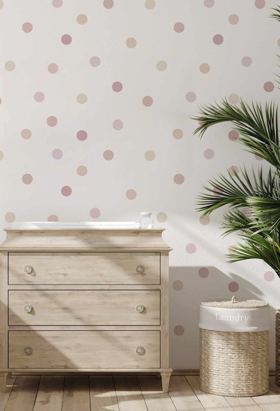 Lifestyle shot of a nursery or child's room featuring a white wall with a repeating pattern of polka dots in varying shades of pink and beige. A light wood dresser with three drawers and round knobs is positioned against the wall, topped with a white changing pad and a small glass jar. A tall green plant with large fronds stands to the right of the dresser, casting shadows on the wall. A woven laundry basket with a white fabric liner embroidered with "Laundry" is on the floor next to the plant.