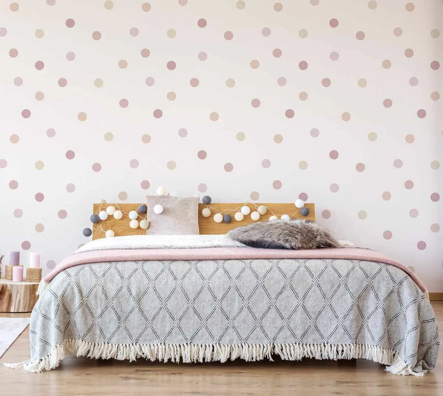 Lifestyle shot of a bedroom featuring Fun Dots 'Melody' wallpaper. The wallpaper has a repeating pattern of irregular polka dots in varying shades of dusty rose, pale pink, and cream on a soft white background. The wallpaper is displayed behind a wooden headboard with a string of decorative white and grey pom-pom lights. The bed is covered with a grey quilted blanket with a fringed edge and a pink throw, with a fluffy grey pillow and a light pink cushion.