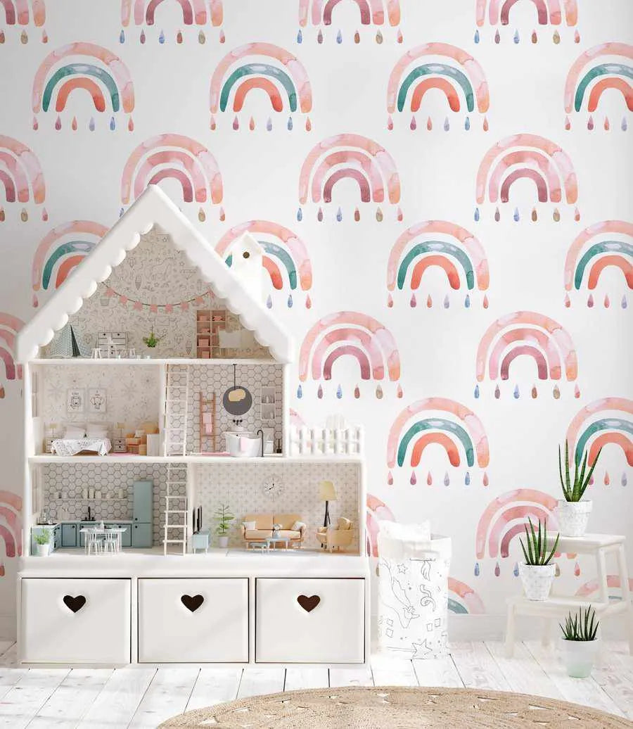 Wallpaper featuring a repeating pattern of hand-painted style rainbows in soft pastel colors including blush pink, dusty rose, sage green, coral, and soft blue, each with matching rain drops below. The design has a watercolor illustration aesthetic on a white background. Shown in a styled children's room with a white wooden dollhouse, white storage boxes with black hearts, potted plants with green foliage, and white flooring, demonstrating how the wallpaper coordinates with soft, neutral nursery decor.