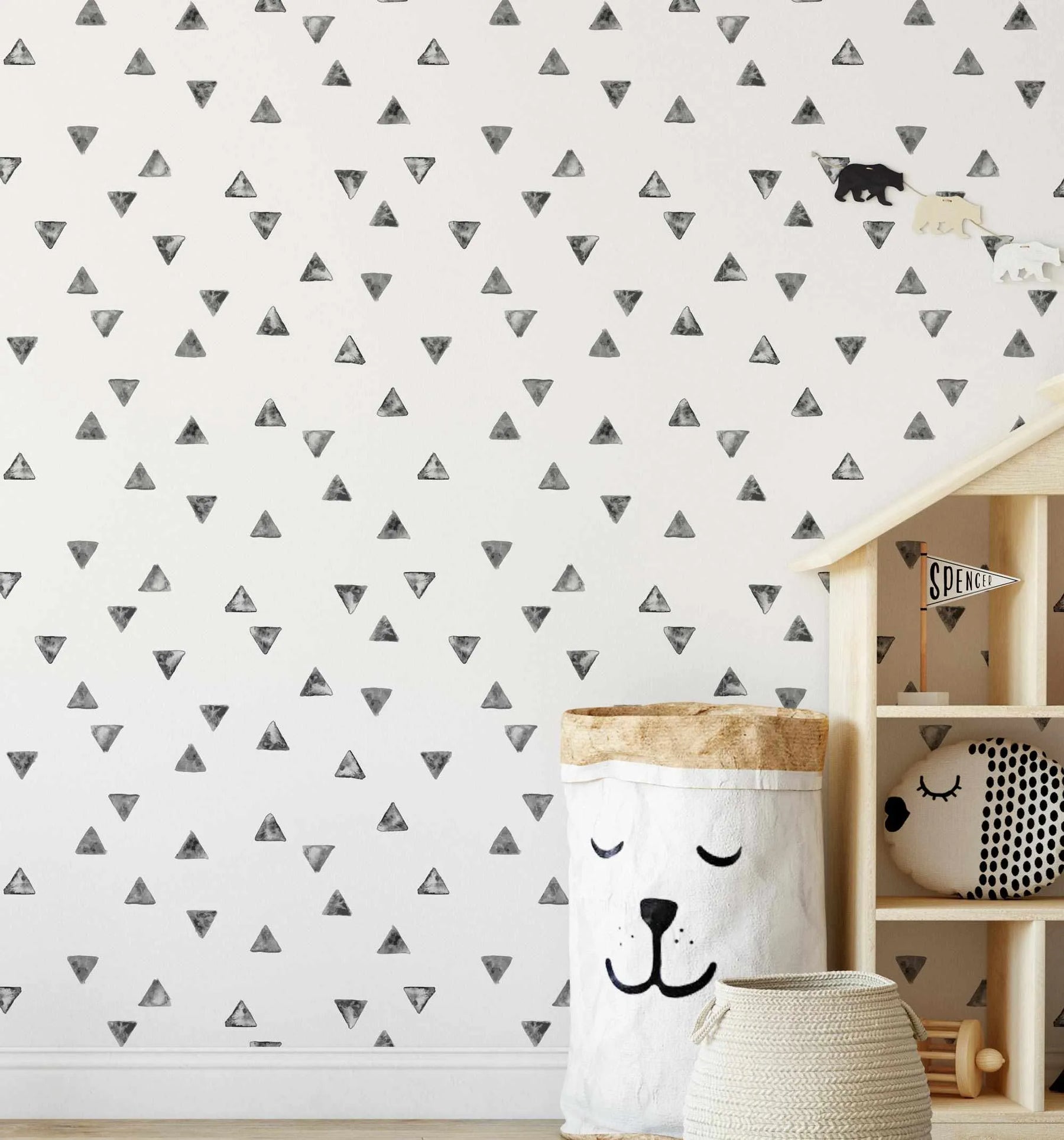 Lifestyle shot of a child's room featuring a white wall with a repeating pattern of hand-painted grey triangles in varying shades and orientations. The room includes a light wood dollhouse-style shelf unit on the right, holding a white pennant flag with "SPENCER" written in black, a black and white spotted pillow, and a wooden toy. A large white fabric storage bin with a bear face design and a smaller woven basket sit on the light wood floor in the foreground.