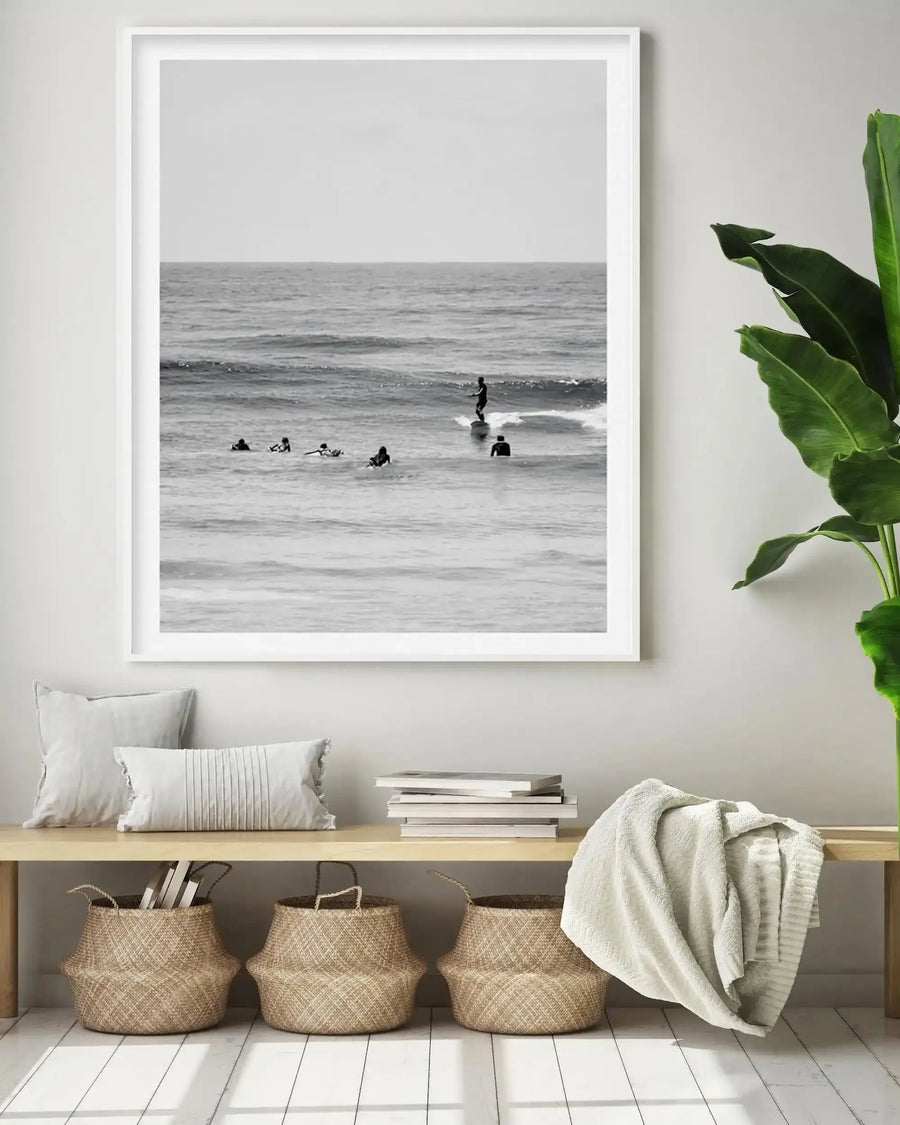Black and white photograph in white frame with white border depicting a calm ocean scene with several surfers in wetsuits scattered across the water, one standing on a surfboard riding a wave. The image is displayed above a wooden console table styled with cream textured pillows, stacked books, three woven natural fiber baskets, and a draped cream linen throw. A green potted plant with large leaves is visible on the right side of the wall.