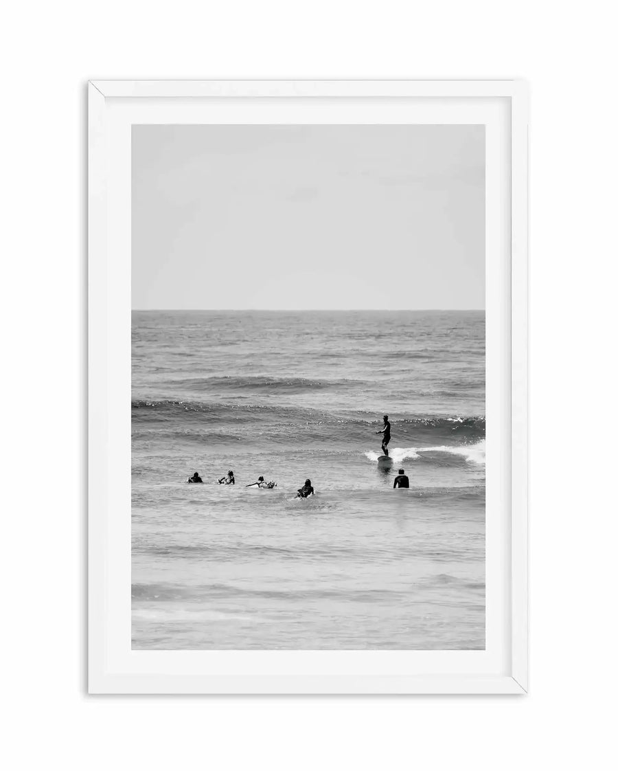 Black and white photograph in white frame with white border depicting several surfers in wetsuits in the ocean. One surfer stands on a wave in the center-right, while others sit on boards in the shallow water to the left. Calm, overcast sky fills the upper portion. Grainy texture and minimalist composition create a serene, contemplative mood of coastal recreation.