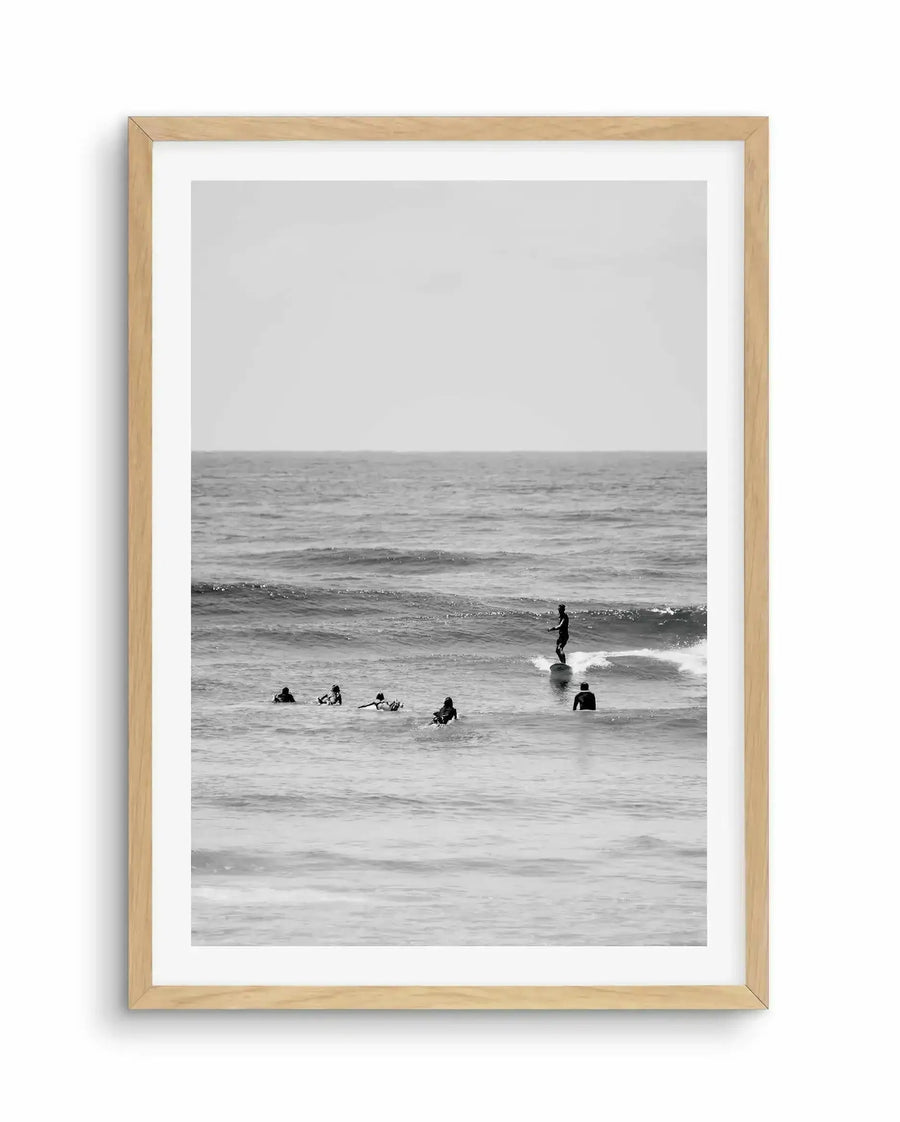 Black and white photograph in natural wood frame with white border showing multiple surfers in wetsuits in the ocean. One surfer stands on a surfboard riding a wave in the center-right, while several others wait in the shallow water. Calm, overcast sky fills the upper half. Soft, muted tones and minimalist composition create a serene, contemplative mood. Photographic print with documentary-style aesthetic.