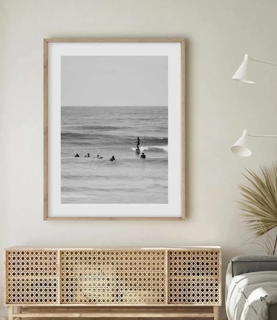 Black and white photograph of surfers and swimmers in ocean waves, displayed in a natural wood frame with white border. The framed print hangs above a light wood dresser with geometric woven panel doors. Two white pendant lamps and a potted plant with green fronds are positioned to the right, with a bed visible in the corner. The photograph captures a beach scene with overcast sky, calm gray-toned water, and multiple figures in the surf, creating a serene coastal atmosphere in a minimalist bedroom setting.