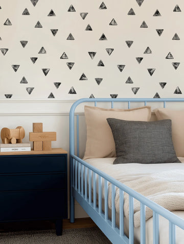 Lifestyle shot of a child's bedroom featuring a light cream wall with a repeating pattern of hand-painted black and grey triangles in various orientations. A light blue metal bed with a white duvet and cream knitted throw is positioned against the wall, with a dark blue bedside table to the left. The table holds two books and wooden toys, including an elephant and building blocks.