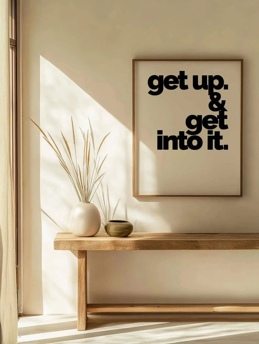 Framed art print displayed on a cream-colored wall above a wooden shelf. The print features bold black sans-serif typography reading "get up. & get into it." on a beige background, housed in a natural wood frame. Below the print, a wooden shelf holds a white ceramic vase with dried grasses and a small brass bowl. Sunlight streams through a window on the left, casting geometric shadows across the wall and shelf. The overall aesthetic is minimalist and warm, with neutral earth tones throughout.
