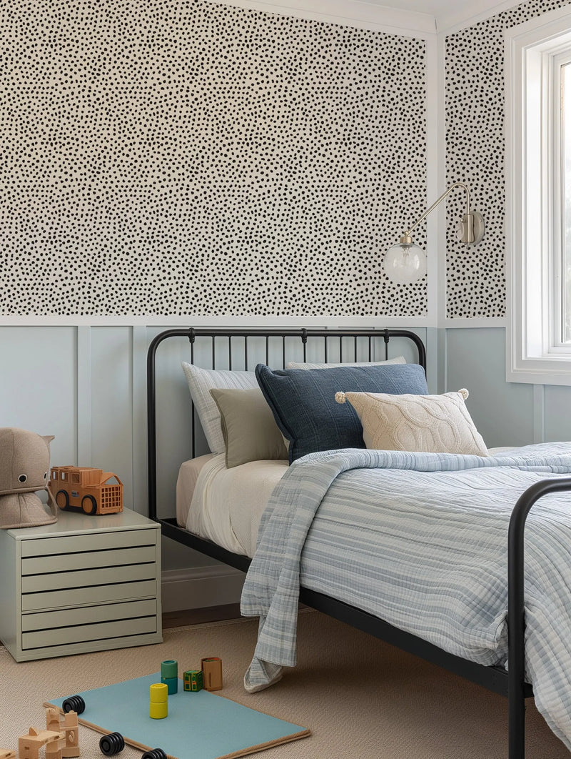 "A child's bedroom featuring Gigi's Dots Wallpaper in Black & Sand on the upper walls, above light blue wainscoting. The wallpaper has a dense, irregular pattern of small black dots on a light sand-colored background. A black metal bed frame with blue and cream bedding is visible, along with a light grey nightstand holding a wooden toy truck and a plush toy. A silver wall sconce with a clear glass globe hangs on the wall, and a window with white trim is on the right."