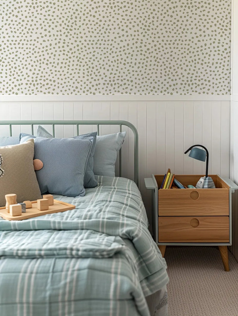 Lifestyle shot of a child's bedroom featuring Gigi's Dots Wallpaper in Khaki on the upper half of the wall above white wainscoting. The wallpaper has a repeating pattern of irregular, organic-shaped dots in a muted khaki green on a light cream background. A bed with a light green metal frame, striped blue and white bedding, and various pillows is visible on the left. A wooden nightstand with two drawers and a blue lamp sits on the right.