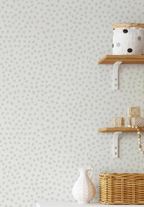 Peel and stick wallpaper featuring a repeating pattern of irregular, hand-drawn dots in a muted sage green on a soft white background. The design has an organic, playful quality. The wallpaper is shown in a room setting with two wooden shelves on the right, displaying a white polka-dot basket, wooden blocks, a beaded garland, a white textured vase, and a woven basket.