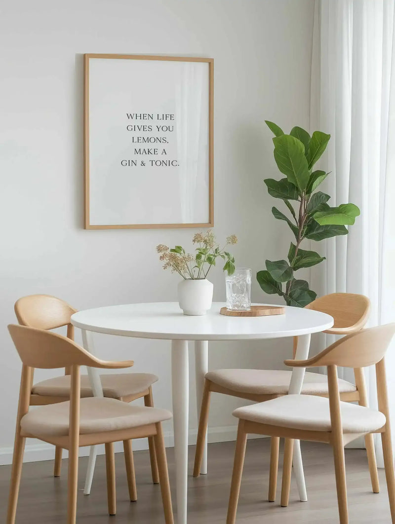 Framed art print with natural wood frame displaying black serif typography on white background reading "WHEN LIFE GIVES YOU LEMONS, MAKE A GIN & TONIC." Shown in a bright, minimalist dining room above a white round table with two natural wood chairs with cream cushions, flanked by a potted fiddle leaf fig plant and small white vase with dried botanicals.