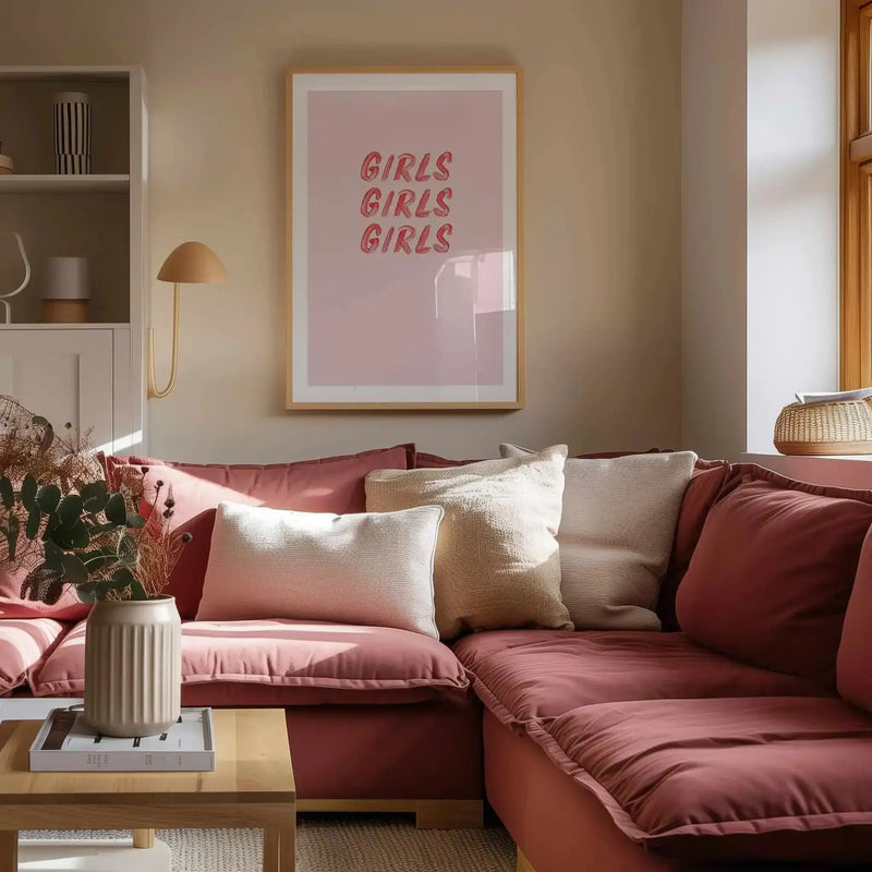 Girls Girls Girls art print displayed in a natural wood frame with white border, hung on a warm beige wall above a burgundy sectional sofa. The print features the phrase "GIRLS GIRLS GIRLS" in bold red italicized lettering centered on a soft mauve-pink background. The framed artwork is shown in a styled living room setting with dusty rose and cream throw pillows, a potted plant with dried flowers in a cream ceramic vase, wooden coffee table, and woven basket accent visible to the right.
