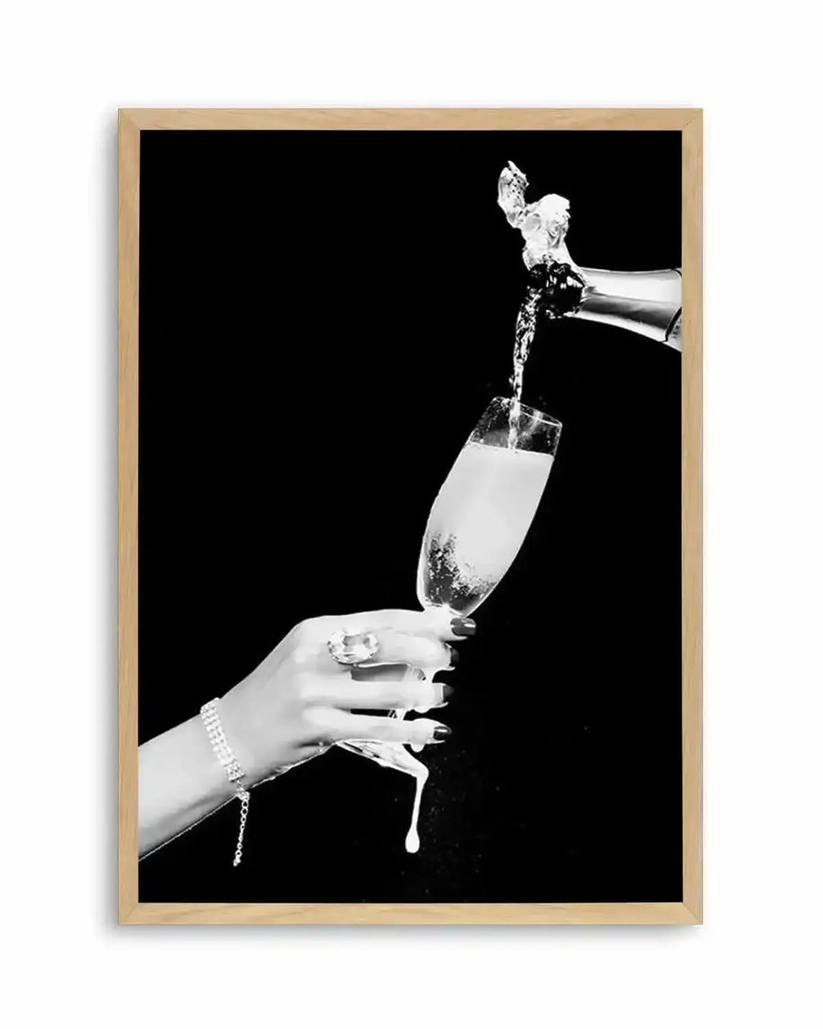 Black and white vintage-style photograph showing a elegant hand holding a champagne flute filled with sparkling champagne being poured from a bottle above. The hand wears a pearl bracelet and has manicured nails. Champagne splashes dramatically from the glass against a solid black background. The artwork is framed in natural wood with a white border.