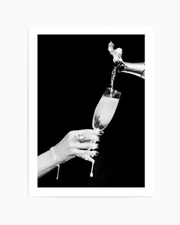 Black and white photograph of a champagne bottle being poured into a flute glass held by a hand wearing a pearl bracelet and ring, with champagne splashing dramatically against a pure black background, printed on white paper with white border.