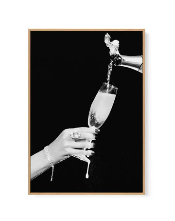 Black and white photograph on canvas print showing a hand with manicured nails and a pearl bracelet holding a champagne flute as champagne is poured from a bottle above it against a solid black background. The canvas is framed in natural wood with a thin border. High contrast monochromatic image with dynamic liquid splash captured mid-pour.