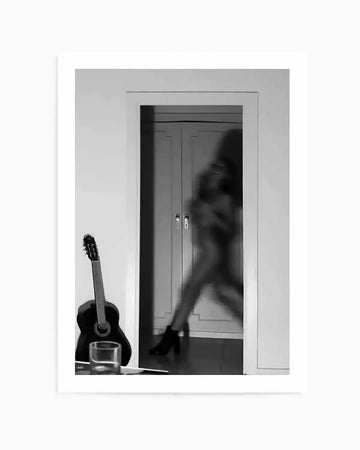 Black and white photograph art print titled "Go Your Own Way" by Mario Stefanelli, showing a blurred silhouette of a person in motion within a doorway, with a classical guitar and glass positioned in the foreground on the left side. The composition uses stark contrast between the white walls and dark interior space, creating a sense of movement and emotion. Displayed in a white frame with white border.