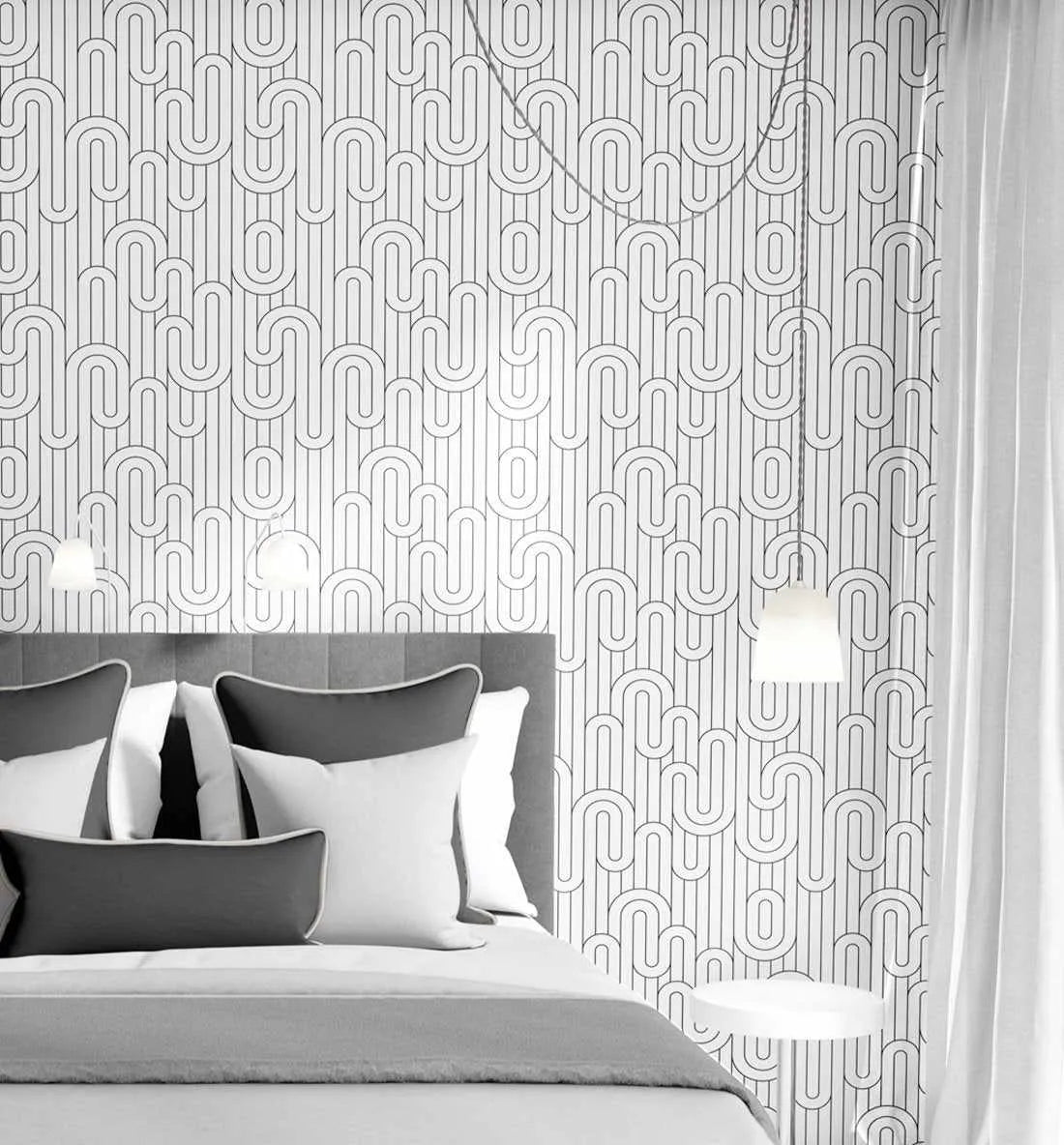 Lifestyle shot of a modern bedroom featuring "Go with the Flow" wallpaper. The wallpaper has a repeating pattern of black lines forming connected U-shapes and vertical stripes on a white background, creating a minimalist, geometric design. The wallpaper covers the wall behind a bed with a grey upholstered headboard, white and grey pillows, and a grey duvet. Two white pendant lights hang from the ceiling, one on each side of the bed, and a white side table is visible on the right.