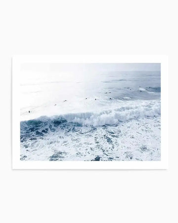 Photography art print featuring an aerial view of surfers in a pale blue ocean with white breaking waves and foamy water. The composition captures the vastness of the ocean under a bright, hazy sky.