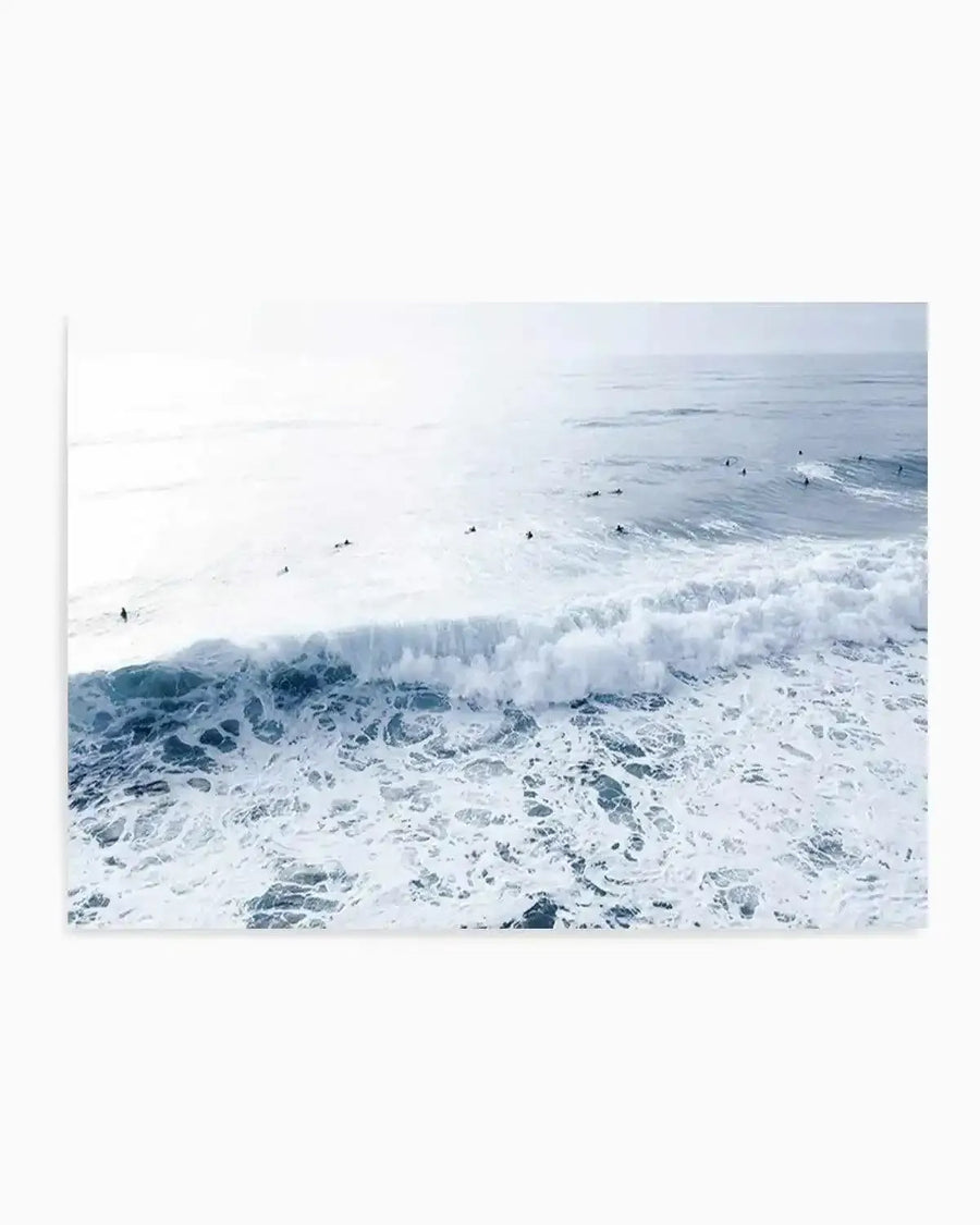 Product shot art print of a coastal photography scene featuring surfers in the ocean. The composition shows a wide expanse of pale blue-grey water under a bright, hazy sky, with a large white breaking wave in the foreground and several smaller waves further out. Numerous surfers are visible as dark silhouettes, some paddling towards the horizon and others waiting for waves. The overall mood is serene and expansive.