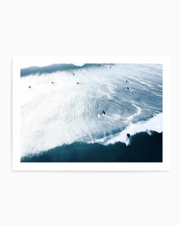 Aerial photograph art print of surfers in a deep blue ocean with white foamy waves. The composition captures multiple surfers scattered across the water, with one surfer actively riding a large breaking wave in the foreground, creating a dynamic and energetic scene.