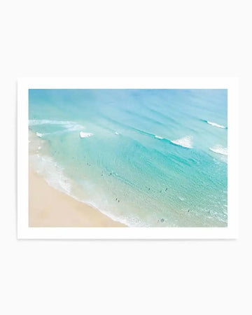 Framed aerial photograph of a beach scene with a natural wood frame and white border. The composition features a light beige sandy beach meeting turquoise and teal ocean water with gentle white waves. Small figures of people are visible in the shallow water and on the sand.