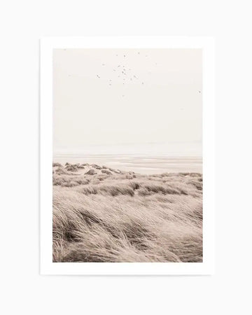 Framed photography art print of a serene coastal landscape with a muted, desaturated colour palette. The foreground features windswept golden-beige dune grasses, leading to gentle sand dunes and a wide, flat beach at low tide. In the distance, the ocean meets a soft, hazy horizon under a pale, almost white sky, with a scattering of small birds flying overhead. The print is displayed with a white border and a simple white frame.