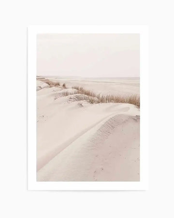 Photography art print featuring a serene coastal landscape with soft, rolling sand dunes in pale beige and dusty rose hues, dotted with sparse, dry golden grasses. The composition extends to a calm, light-coloured ocean under a hazy, bright sky, creating a tranquil, minimalist mood.